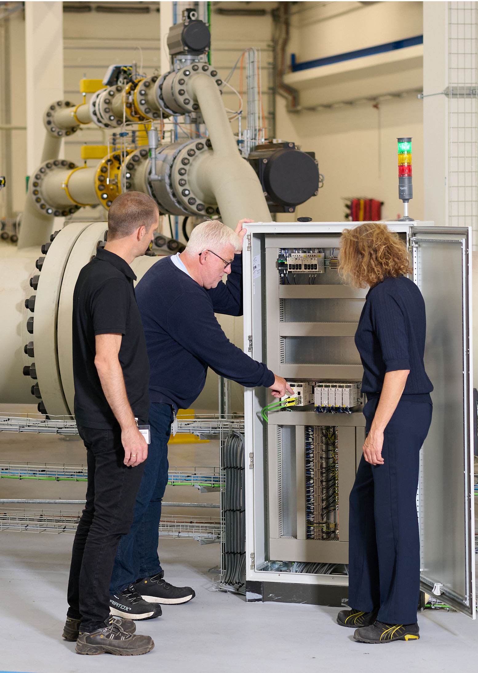 Three persons around a PLC cabinet with green light on top. There are big pipes in the background.