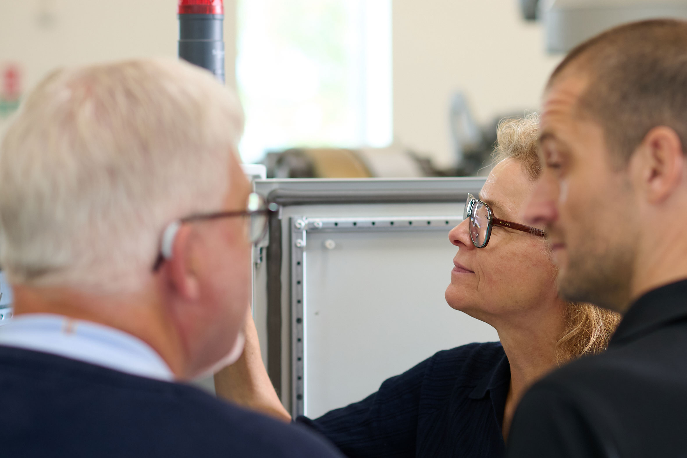 To men with their backs to the camera facing slightly sideway looking at each other. Behind them a woman is looking at screen.