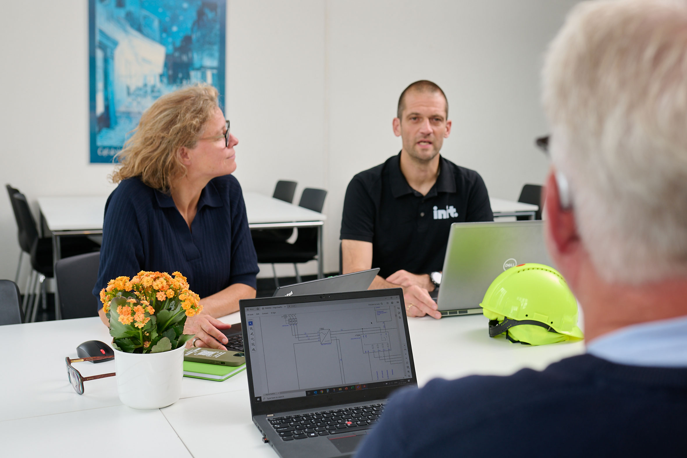 Three people sitting around a table. One has his bag to the camera and you can see a flow chart on hi laptop screen. There is a hard hat and a pot of flowers on the table.