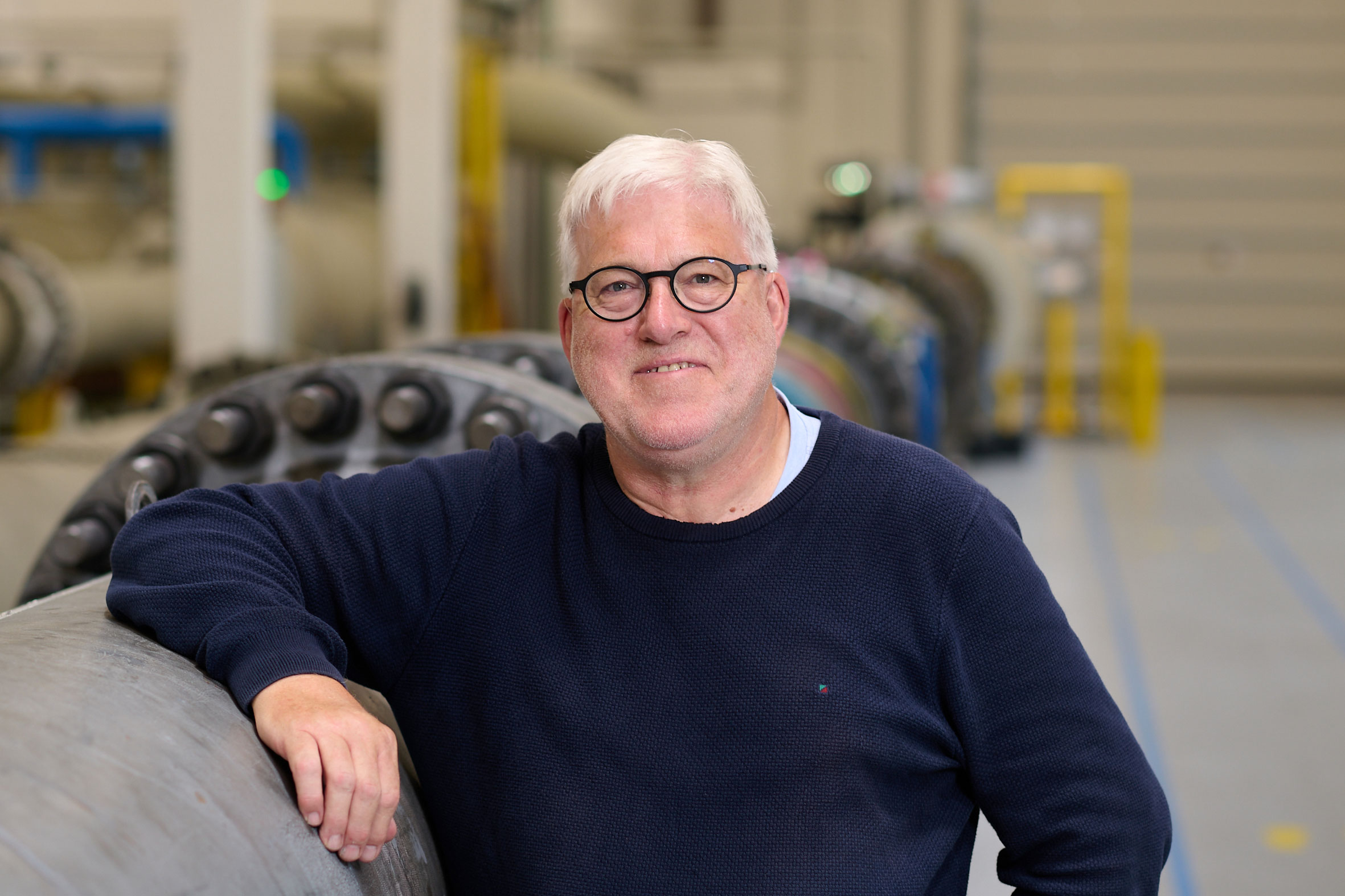 Man with white hair and round glasses, wearing a dark blue sweater is leaning on a large pipe and looking at the camera.