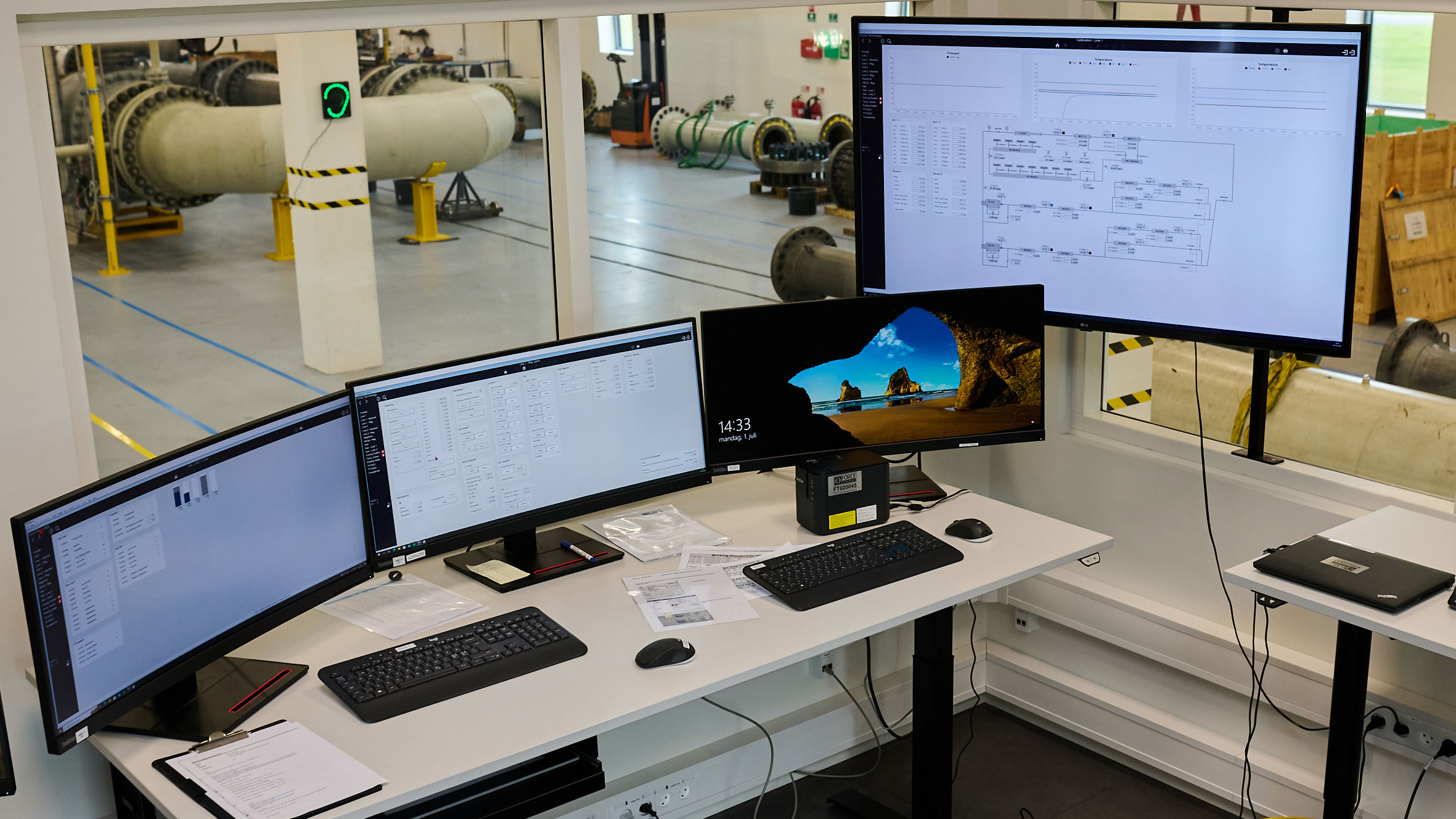 Three small computer screens and one large screen placed in a factory setting with big pipes.